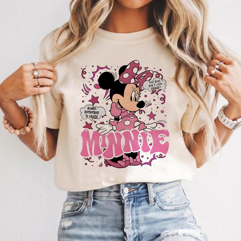 Op de afbeelding: Cr&egrave;mekleurig T-shirt met een afbeelding van Minnie Mouse in een roze jurk met stippen en een strik. Het woord "MINNIE" staat in roze, gestileerde letters. Tekstballonnen zeggen "Always Remember To Laugh" en "Too-Cool Ain't Life Grand?"