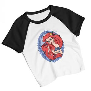 May include: White raglan t-shirt with black sleeves and collar. The shirt features a cartoon illustration of a mermaid with red hair, a blue seashell bra, and pink flowers, set within a blue oval frame.