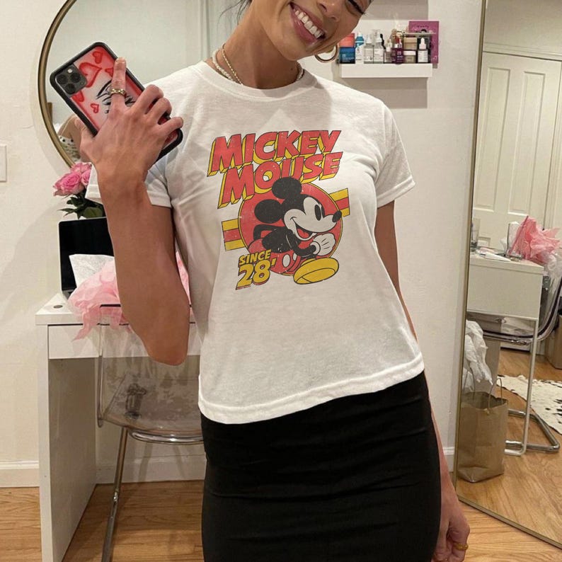 May include: White t-shirt with a Mickey Mouse graphic. The design features the words "Mickey Mouse" in red and yellow, with Mickey's image in the center. The text "Since 28" is also visible. The person is wearing a black skirt.