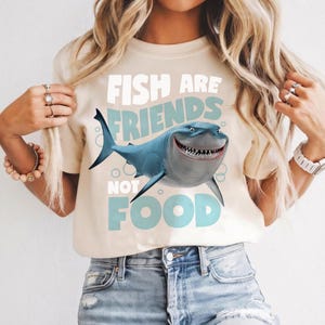 Op de afbeelding: Beige T-shirt met een cartoon haai. De blauwe haai heeft een brede grijns en scherpe tanden. De tekst "FISH ARE FRIENDS NOT FOOD" staat in lichtblauw boven en onder de haai.