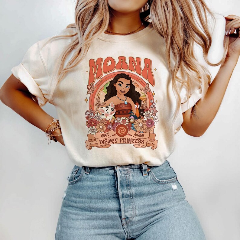 May include: A cream-colored short-sleeve t-shirt featuring a graphic print of Moana, Pua, and Heihei surrounded by flowers and butterflies. The text reads MOANA at the top and EST 2016 DISNEY PRINCESS at the bottom in a retro orange font.