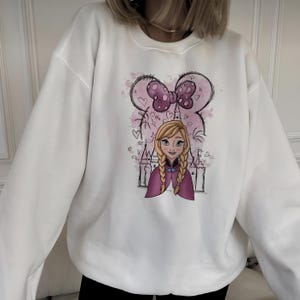 May include: A white crewneck sweatshirt featuring a graphic print of a blonde character with braided hair, wearing a purple cape. The design includes a pink watercolor background, a castle outline, and a large purple bow with polka dots above.