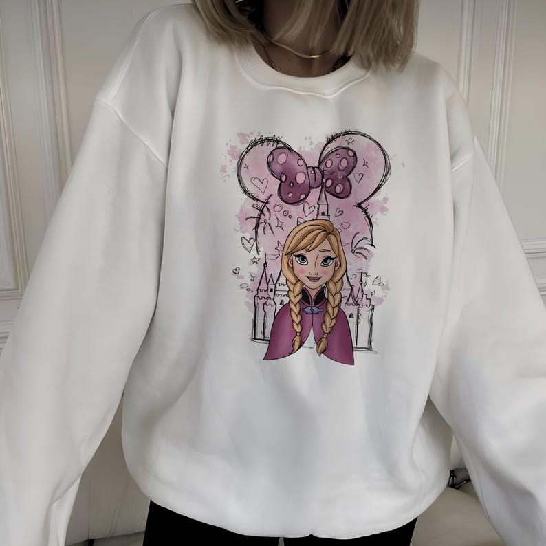 May include: A white crewneck sweatshirt featuring a graphic print of a blonde character with braided hair, wearing a purple cape. The design includes a pink watercolor background, a castle outline, and a large purple bow with polka dots above.