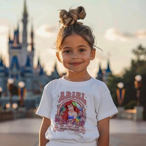 May include: A white short-sleeved t-shirt featuring a graphic print of Ariel with red hair, surrounded by colourful flowers, butterflies, and the text Ariel, Est 1989, Disney Princess. The shirt has a crew neckline and a straight hemline.