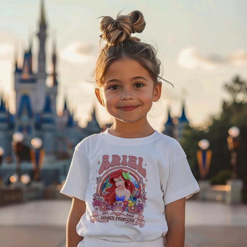 May include: A white short-sleeved t-shirt featuring a graphic print of Ariel with red hair, surrounded by colourful flowers, butterflies, and the text Ariel, Est 1989, Disney Princess. The shirt has a crew neckline and a straight hemline.