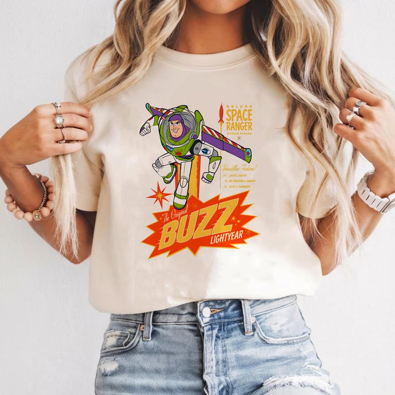 May include: Cream-colored t-shirt featuring a graphic of Buzz Lightyear in a dynamic pose. The design includes the text "The Original BUZZ LIGHTYEAR" in a red and yellow burst, and "DELUXE SPACE RANGER" with additional text. The shirt has short sleeves.