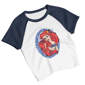 May include: White raglan t-shirt with navy blue sleeves and collar. The shirt features a cartoon illustration of a mermaid with red hair, a blue seashell bra, and pink flowers, framed in a blue oval.