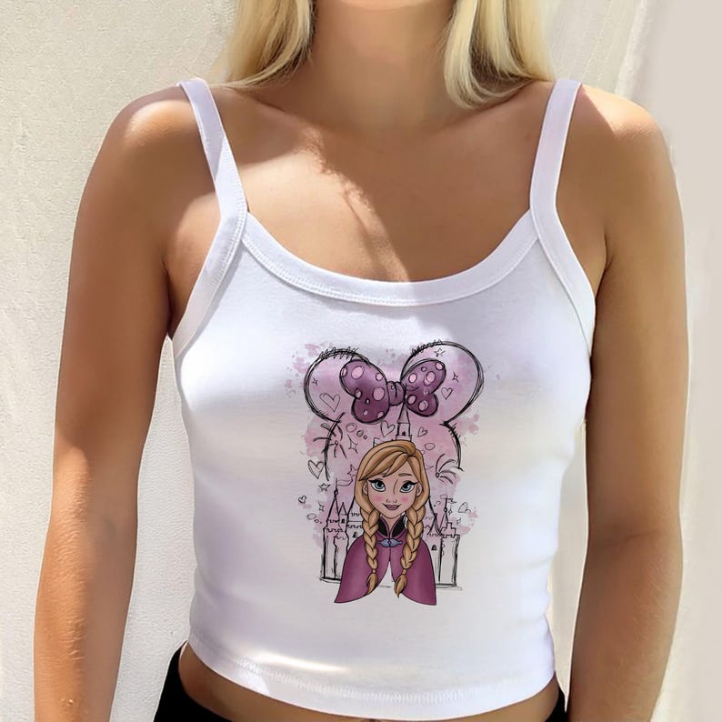 May include: A white sleeveless tank top featuring a graphic print of a cartoon character with blonde braided hair, wearing a purple cape. Behind the character is a purple polka-dot bow, a castle silhouette, and small heart and star outlines.