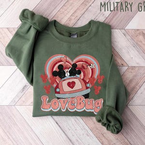Pode incluir: Camisola verde militar com um gráfico com um carro com Mickey e Minnie Mouse, um arco-íris em forma de coração e o texto "LoveBug". O design inclui corações vermelhos e uma flor. A camisola é exibida numa superfície de madeira.