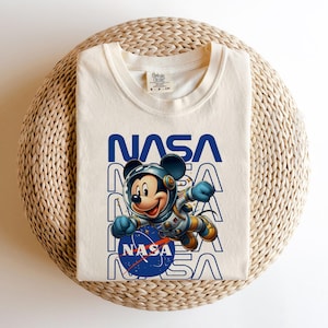 May include: Cream-colored t-shirt featuring a graphic of Mickey Mouse in a spacesuit, with the NASA logo and text. The design is in blue and white. The shirt is folded on a woven, light brown surface.