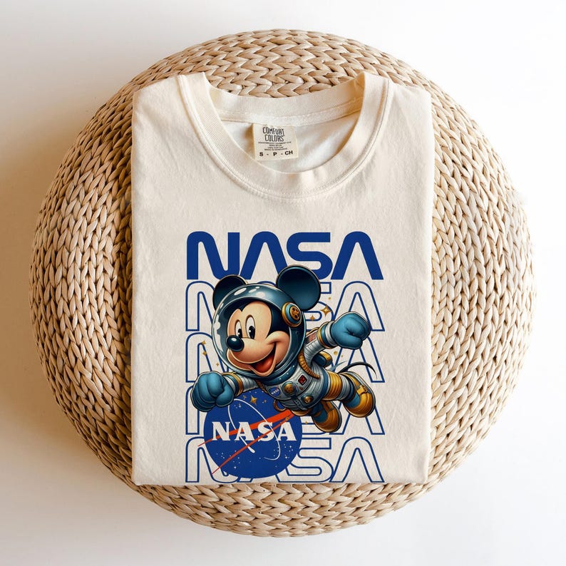 May include: Cream-colored t-shirt featuring a graphic of Mickey Mouse in a spacesuit, with the NASA logo and text. The design is in blue and white. The shirt is folded on a woven, light brown surface.