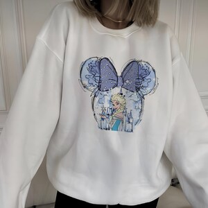 May include: White sweatshirt featuring a blue and white graphic of Elsa from Frozen inside a Mickey Mouse head shape. The design includes a blue bow with white polka dots and a castle. The sweatshirt is a casual style.