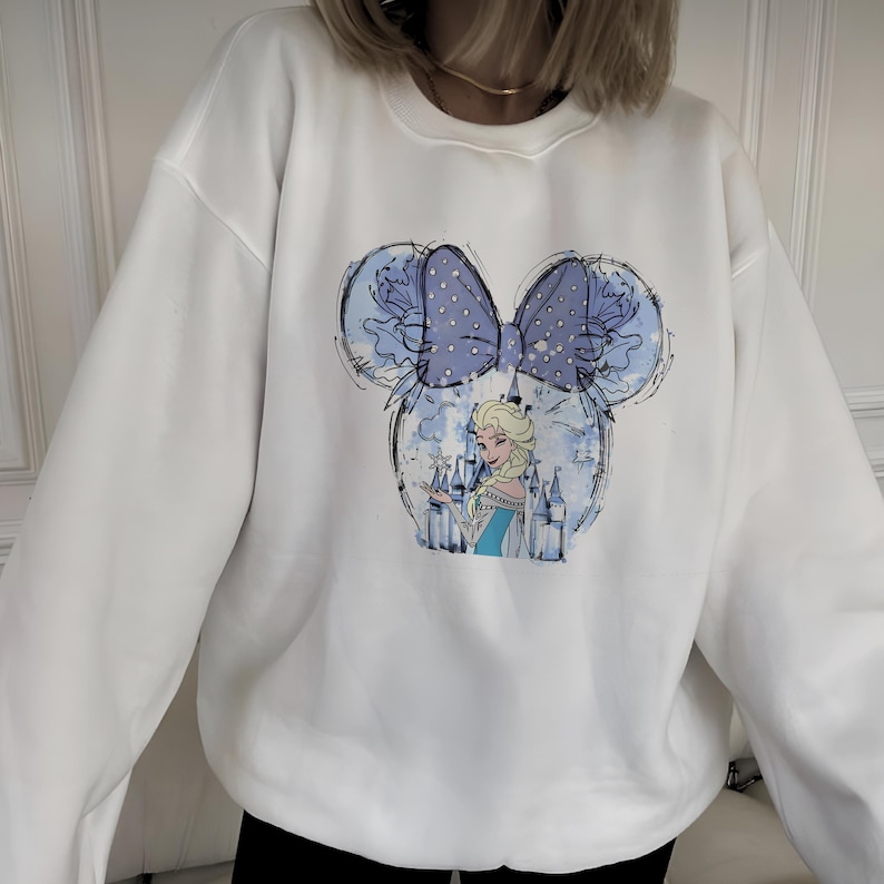 May include: White sweatshirt featuring a blue and white graphic of Elsa from Frozen inside a Mickey Mouse head shape. The design includes a blue bow with white polka dots and a castle. The sweatshirt is a casual style.