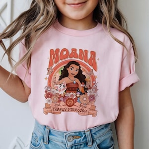 May include: A light pink t-shirt featuring a graphic print of Moana, Pua the pig, and Heihei the rooster. The design includes orange and brown floral accents, butterflies, and the text "MOANA EST 2016 DISNEY PRINCESS" in a retro-style font.