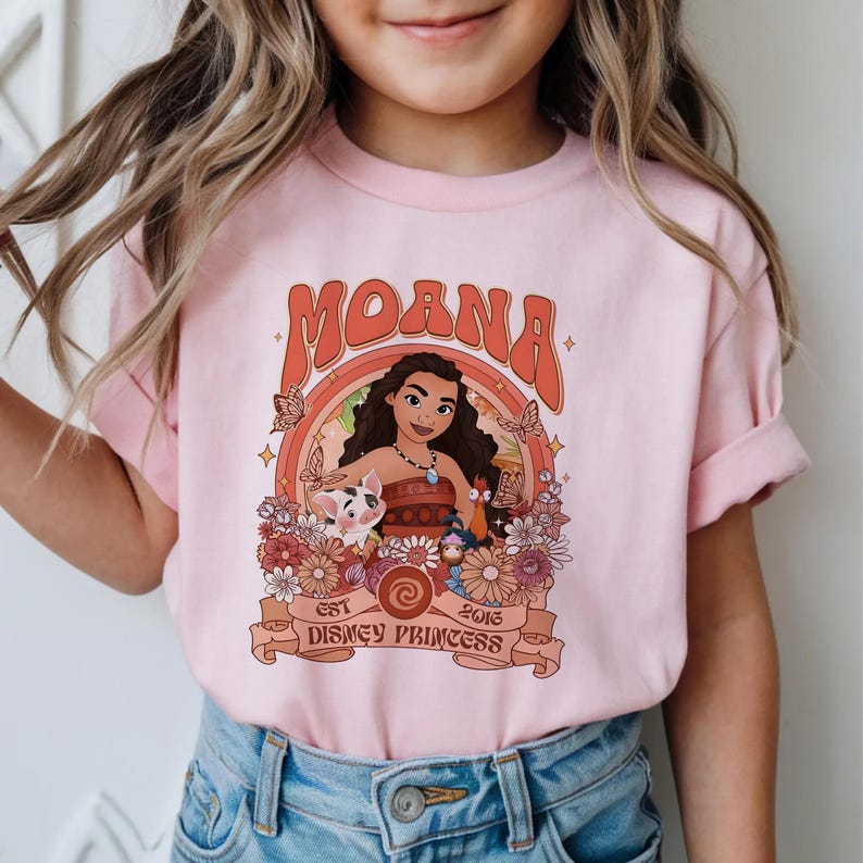 May include: A light pink t-shirt featuring a graphic print of Moana, Pua the pig, and Heihei the rooster. The design includes orange and brown floral accents, butterflies, and the text "MOANA EST 2016 DISNEY PRINCESS" in a retro-style font.