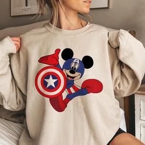 Peut inclure: Sweat-shirt beige avec Mickey Mouse en Captain America. Mickey porte un casque bleu avec un "A" blanc, des gants et des bottes rouges, et tient un bouclier avec une &eacute;toile blanche. Le design est rouge, blanc et bleu.