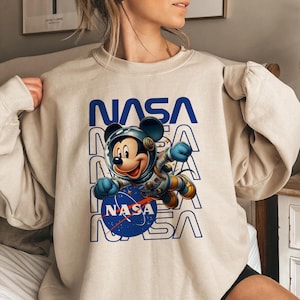 May include: Beige sweatshirt featuring a graphic of Mickey Mouse in a NASA spacesuit. The word "NASA" is printed in blue above and below the character. The NASA logo is on the blue circle. The sweatshirt is a casual style.