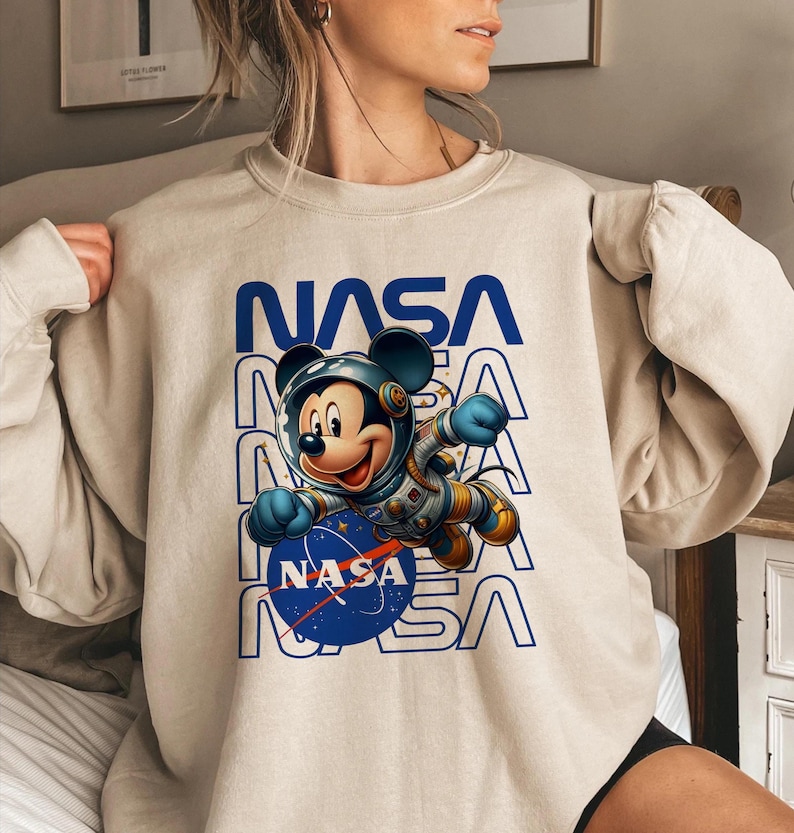 May include: Beige sweatshirt featuring a graphic of Mickey Mouse in a NASA spacesuit. The word "NASA" is printed in blue above and below the character. The NASA logo is on the blue circle. The sweatshirt is a casual style.