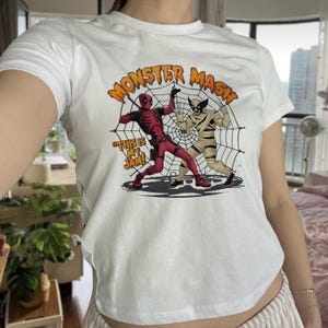 May include: A white short-sleeve t-shirt featuring a graphic print of Deadpool and Wolverine dressed as a mummy. The text reads "MONSTER MASH!" and "THIS IS MY JAM!" above and beside the characters, who are positioned in front of a spider web.