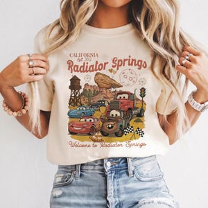 Pode incluir: Camiseta bege com um gr&aacute;fico colorido de carros de desenho animado e o texto "California Est. 2012 Radiator Springs". O design inclui uma paisagem des&eacute;rtica, uma torre de &aacute;gua e a frase "Welcome to Radiator Springs".
