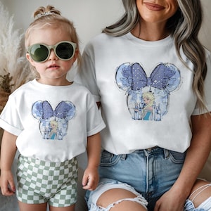 May include: White t-shirts featuring a blue and purple Minnie Mouse design with a princess and castle. The design includes a polka dot bow and floral accents. One shirt is on a child, the other on an adult.