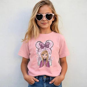 May include: A light pink short-sleeved t-shirt featuring a graphic print of a cartoon character with blonde braided hair, a purple cape, and a castle silhouette background, topped with a large purple polka-dot bow and heart outlines.