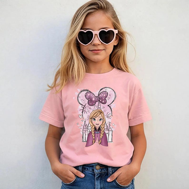 May include: A light pink short-sleeved t-shirt featuring a graphic print of a cartoon character with blonde braided hair, a purple cape, and a castle silhouette background, topped with a large purple polka-dot bow and heart outlines.