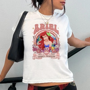 K&ouml;nnte beinhalten: Ein wei&szlig;es Kurzarm-T-Shirt mit einer Grafik von Arielle, die Meerjungfrau, mit rotem Haar, umgeben von Blumen, Schmetterlingen und Fabius. Der Text lautet ARIEL, EST 1989 und DISNEY PRINCESS, eingerahmt in einem kreisf&ouml;rmigen rosa Rand.