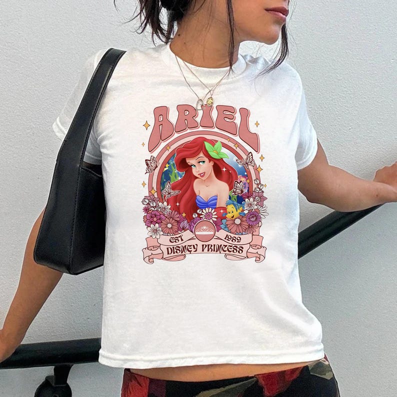 K&ouml;nnte beinhalten: Ein wei&szlig;es Kurzarm-T-Shirt mit einer Grafik von Arielle, die Meerjungfrau, mit rotem Haar, umgeben von Blumen, Schmetterlingen und Fabius. Der Text lautet ARIEL, EST 1989 und DISNEY PRINCESS, eingerahmt in einem kreisf&ouml;rmigen rosa Rand.