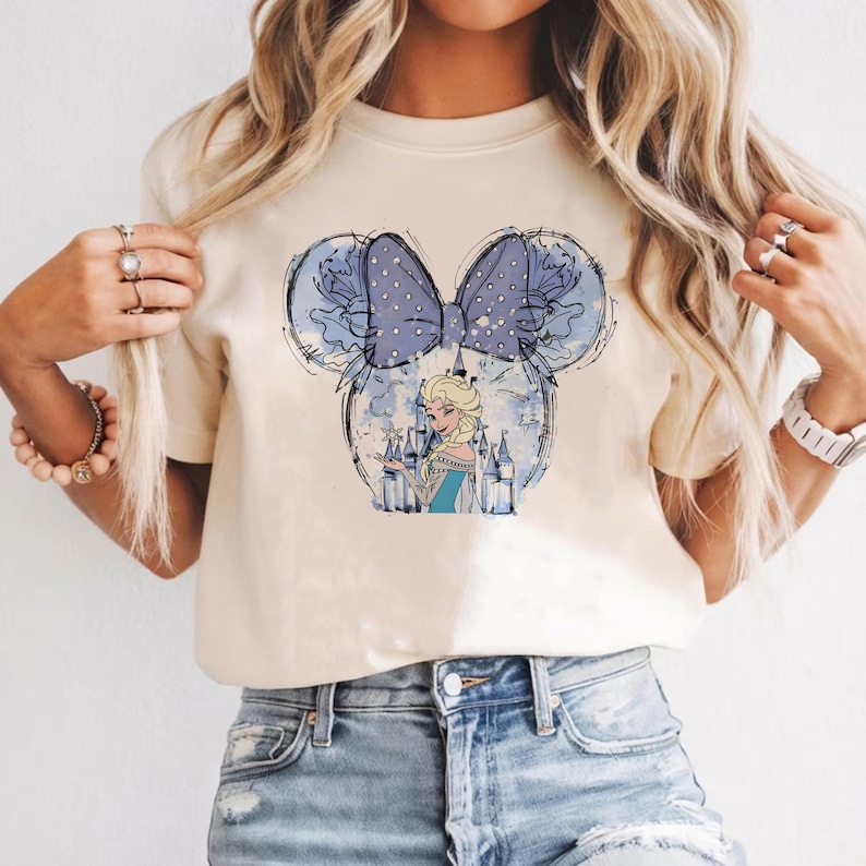 May include: Cream-colored t-shirt featuring a graphic of a cartoon character in a castle, inside a Mickey Mouse ear design. The ears are outlined in black with a blue bow with white polka dots. The character is wearing a blue dress.