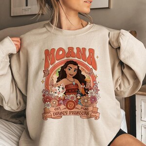 May include: A beige crewneck sweatshirt featuring a graphic print of the character Moana, a pig, and a rooster surrounded by flowers and butterflies. The text reads MOANA, EST 2016, and DISNEY PRINCESS, with a circular frame and orange accents.