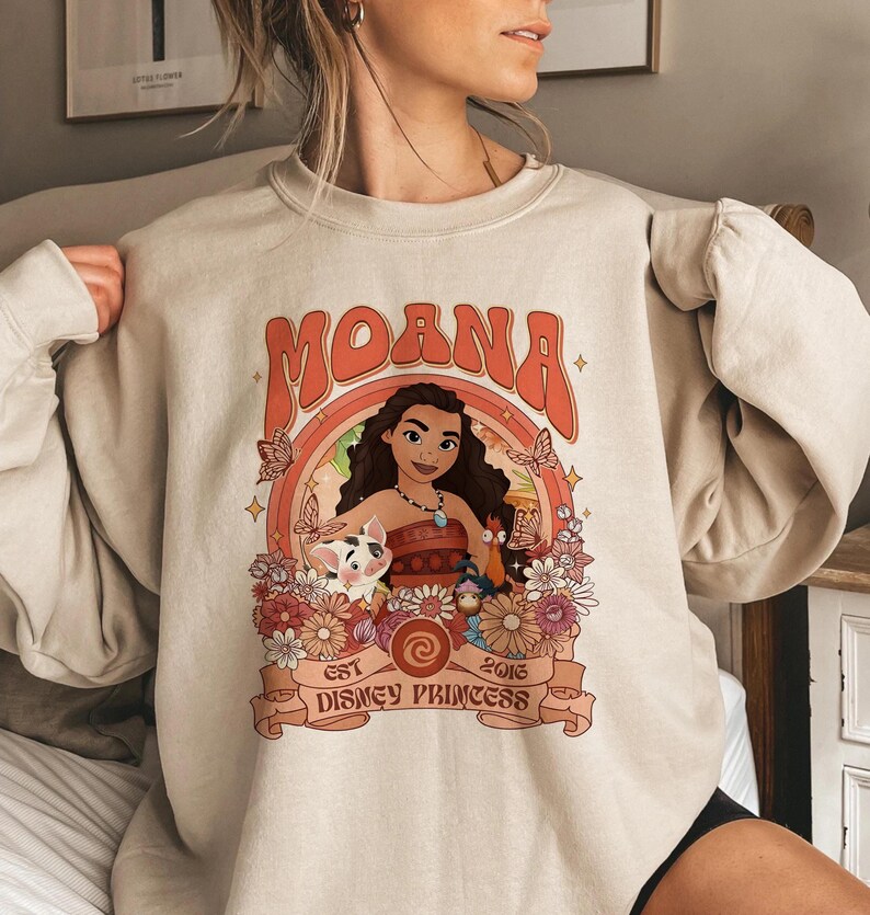 May include: A beige crewneck sweatshirt featuring a graphic print of the character Moana, a pig, and a rooster surrounded by flowers and butterflies. The text reads MOANA, EST 2016, and DISNEY PRINCESS, with a circular frame and orange accents.