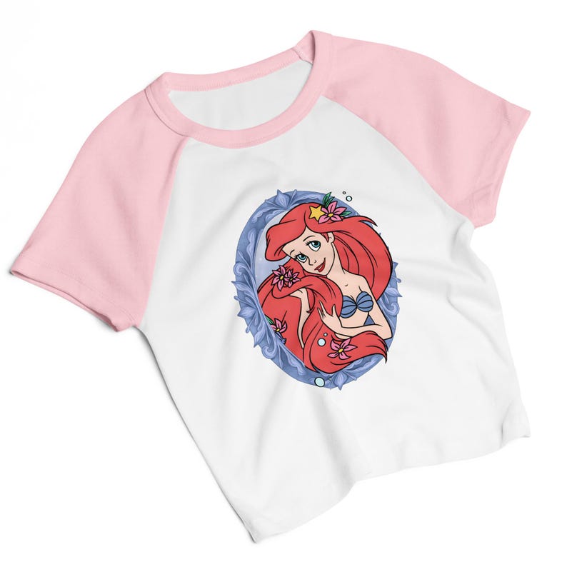May include: White raglan-sleeve t-shirt with pink sleeves and a crew neck. The front features a cartoon illustration of a red-haired mermaid in a blue oval frame, wearing a blue seashell bra and holding flowers.