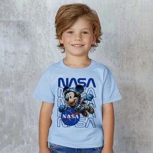 May include: A light blue t-shirt featuring Mickey Mouse in a NASA spacesuit. The word "NASA" is printed in blue vertically on the shirt. The image shows a young child wearing the shirt.