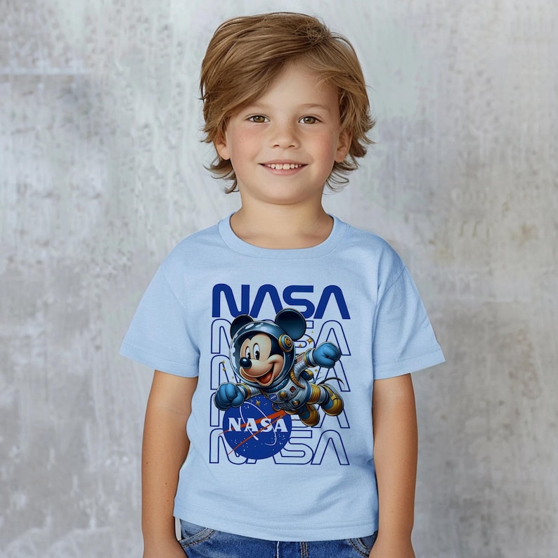 May include: A light blue t-shirt featuring Mickey Mouse in a NASA spacesuit. The word "NASA" is printed in blue vertically on the shirt. The image shows a young child wearing the shirt.