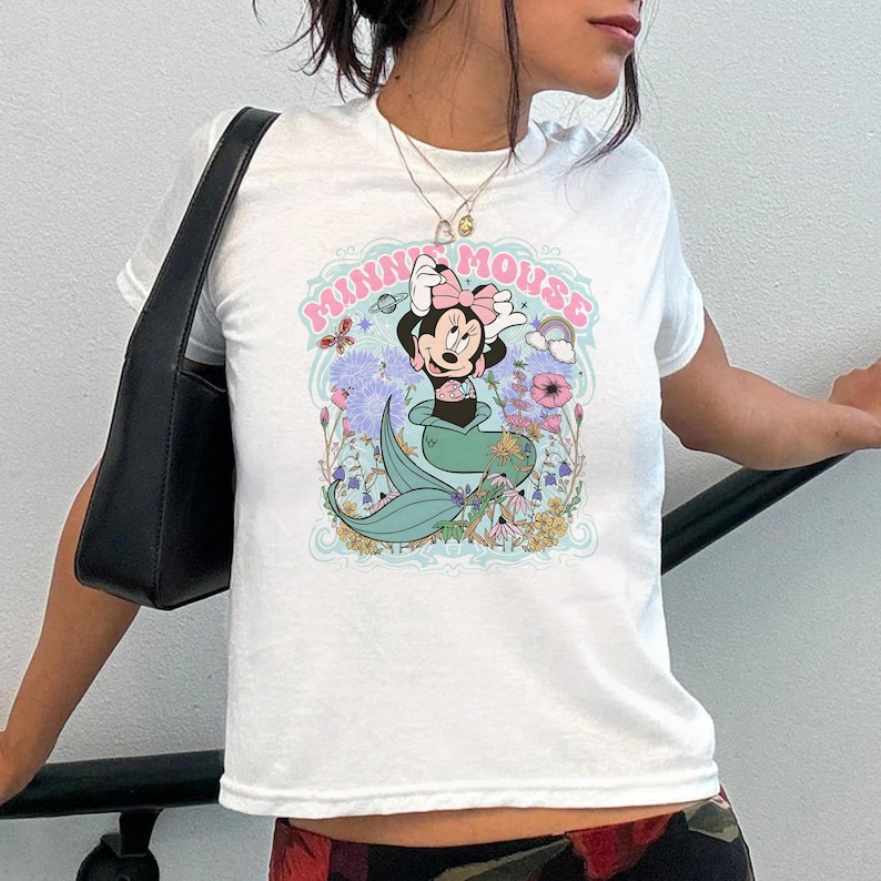 Op de afbeelding: Wit T-shirt met een Minnie Mouse zeemeermin ontwerp. De afbeelding toont Minnie met een roze strik en een turquoise staart, met de woorden "MINNIE MOUSE" in het roze. Het ontwerp is omgeven door bloemen en vlinders.
