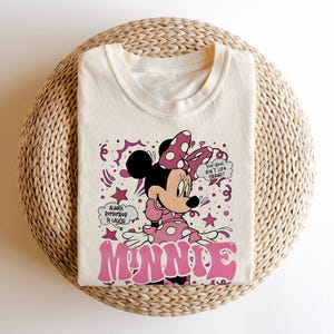 Op de afbeelding: Cr&egrave;mekleurig T-shirt met een afbeelding van Minnie Mouse in een roze polkadot jurk en strik. Het woord "MINNIE" staat in roze bubbelletters. Tekst bevat "Always remember to laugh" en "Too-Yoo! Ain't life grand?"