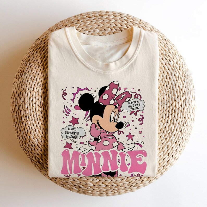 Op de afbeelding: Cr&egrave;mekleurig T-shirt met een afbeelding van Minnie Mouse in een roze polkadot jurk en strik. Het woord "MINNIE" staat in roze bubbelletters. Tekst bevat "Always remember to laugh" en "Too-Yoo! Ain't life grand?"
