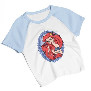 May include: White and light blue raglan t-shirt featuring a cartoon illustration of a red-haired mermaid in a blue oval frame. The mermaid has a blue seashell bra and is adorned with pink flowers and a yellow star.