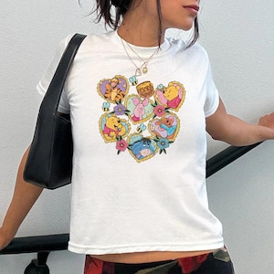 May include: White t-shirt featuring a colorful heart-shaped design with cartoon characters. The design includes Winnie the Pooh, Piglet, Tigger, Eeyore, and bees. The shirt has a crew neck and short sleeves.