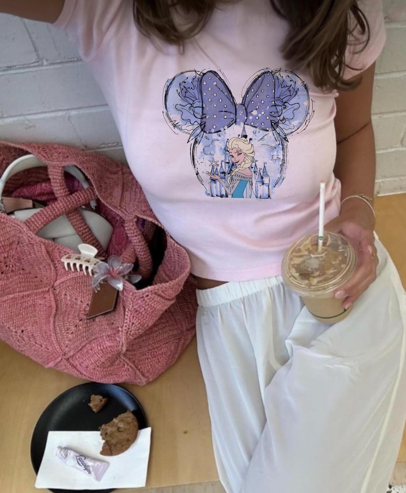 May include: A light pink t-shirt featuring a Minnie Mouse-inspired design with a blue bow and an illustration of a princess. A pink knitted tote bag, a black plate with a cookie, and a clear cup with a straw are also visible.