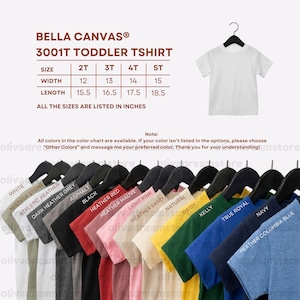 Puede incluir: Una camiseta blanca para ni&ntilde;os peque&ntilde;os colgada de una percha negra. La imagen incluye una carta de colores con varios colores de camisetas y una tabla de tallas en pulgadas para las tallas 2T a 5T. El texto dice "BELLA CANVAS 3001T TODDLER TSHIRT."