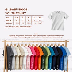 Puede incluir: Exhibici&oacute;n de camisetas Gildan 5000B para j&oacute;venes en varios colores, colgadas en un perchero de madera. La tabla de tallas muestra anchos de 40.6 cm a 50.8 cm y largos de 52.1 cm a 67.3 cm. Colores: carb&oacute;n, ceniza, natural, blanco, etc.