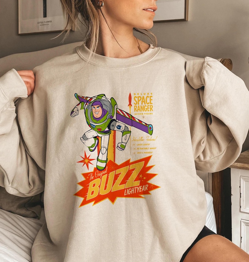 May include: Beige sweatshirt featuring a graphic of Buzz Lightyear in his space ranger suit. The design includes the text "The Original Buzz Lightyear" in a red and yellow starburst, and "Deluxe Space Ranger" above the character.