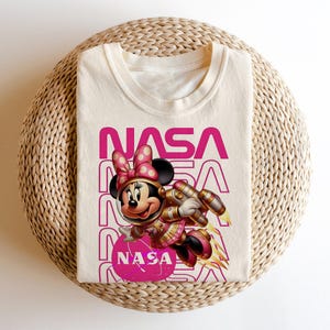 May include: Cream t-shirt with a cartoon Minnie Mouse in a space suit, and the word "NASA" in pink. Minnie Mouse has a polka-dot bow and a space gun. The shirt is folded on a woven surface.