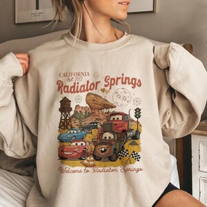 Pode incluir: Camisola bege com um gr&aacute;fico de Radiator Springs do filme Carros. O design inclui personagens de carros, uma paisagem des&eacute;rtica e o texto "CALIFORNIA est. 2012 Radiator Springs" e "Welcome to Radiator Springs".