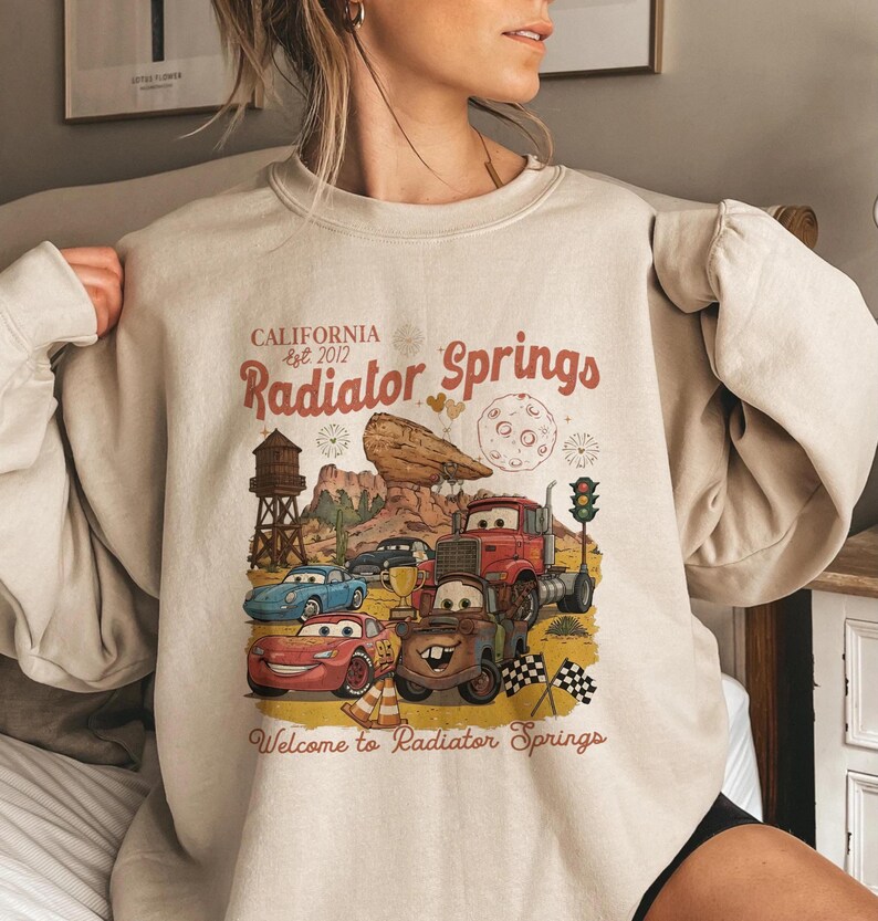 Pode incluir: Camisola bege com um gr&aacute;fico de Radiator Springs do filme Carros. O design inclui personagens de carros, uma paisagem des&eacute;rtica e o texto "CALIFORNIA est. 2012 Radiator Springs" e "Welcome to Radiator Springs".
