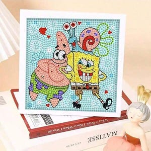 May include: A square diamond painting featuring the cartoon characters SpongeBob SquarePants and Patrick Star. The artwork is set against a light blue background with a border of small, colorful gems. The characters are depicted in their signature outfits.
