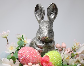 May include: A silver bunny figurine sits amidst a colorful arrangement of flowers and decorative eggs. The Easter-themed display features pink, green, and white eggs, with delicate blossoms and greenery. The bunny has long ears and is the focal point.