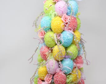 May include: A colorful Easter egg tree decoration. The tree is composed of many pastel-colored, glitter-covered eggs, including yellow, pink, blue, and green. It is adorned with pink roses, ribbons, and decorative sprigs, and topped with bunny ears.
